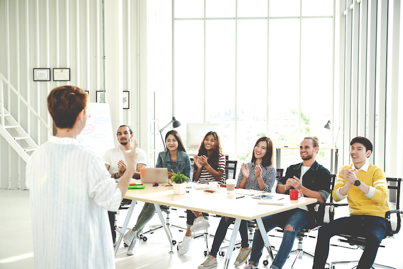 Businesswoman explain ideas to group of creative diverse team at modern office. Rear view of manager gesturing hand standing against multiethnic people. Audience applauding speaker after presentation.